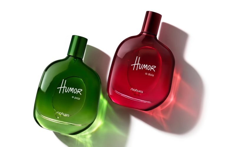 Foto com fundo branco. Dois frascos de perfume Humor Paz e Humor em verde e Humor a Dois em vermelho, sobre superfície refletiva.